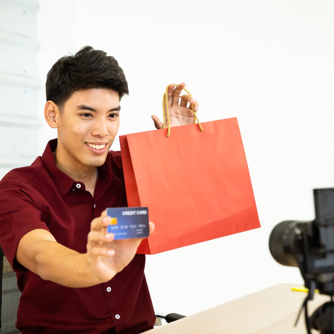 Young Asian male online merchant blogger using camera to live vi Young Asian male online merchant blogger using camera to live video, show shopping bag and credit card. Social media, Influencer, online shopping, and payment concept.